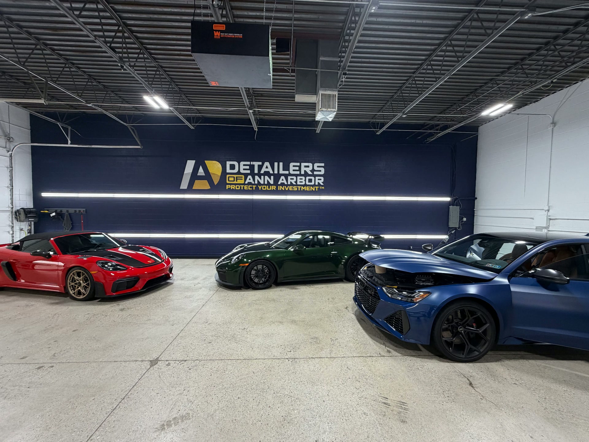 Porsches and an Audi R8 staged in front of the Detailers of Ann Arbor logo wall